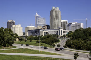 City skyline with tall buildings behind a grassy foreground and a curving multi-lane road in clear weather.